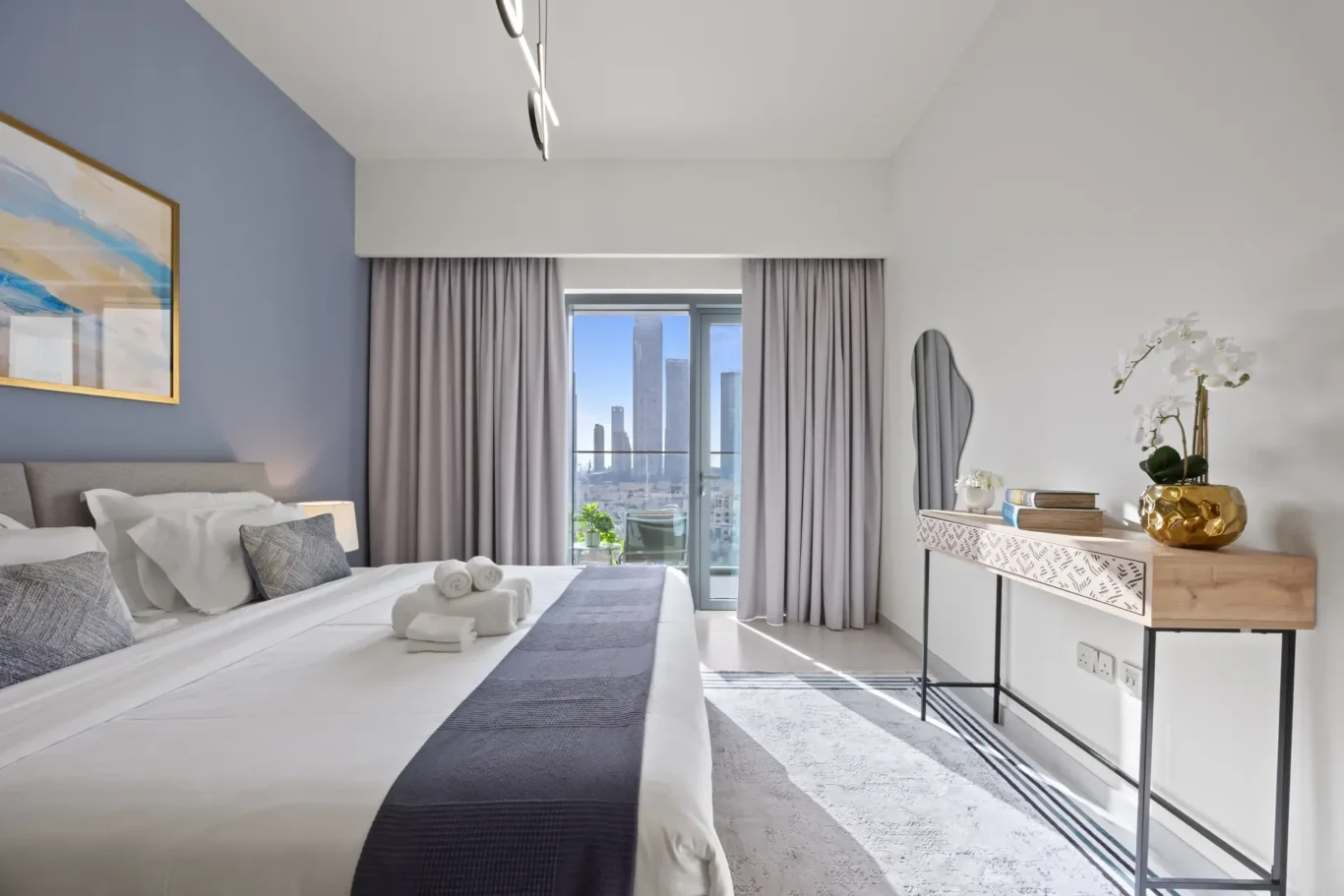Hotel bedroom with two beds, a blue accent wall, and a balcony view of a city skyline through tall curtains.