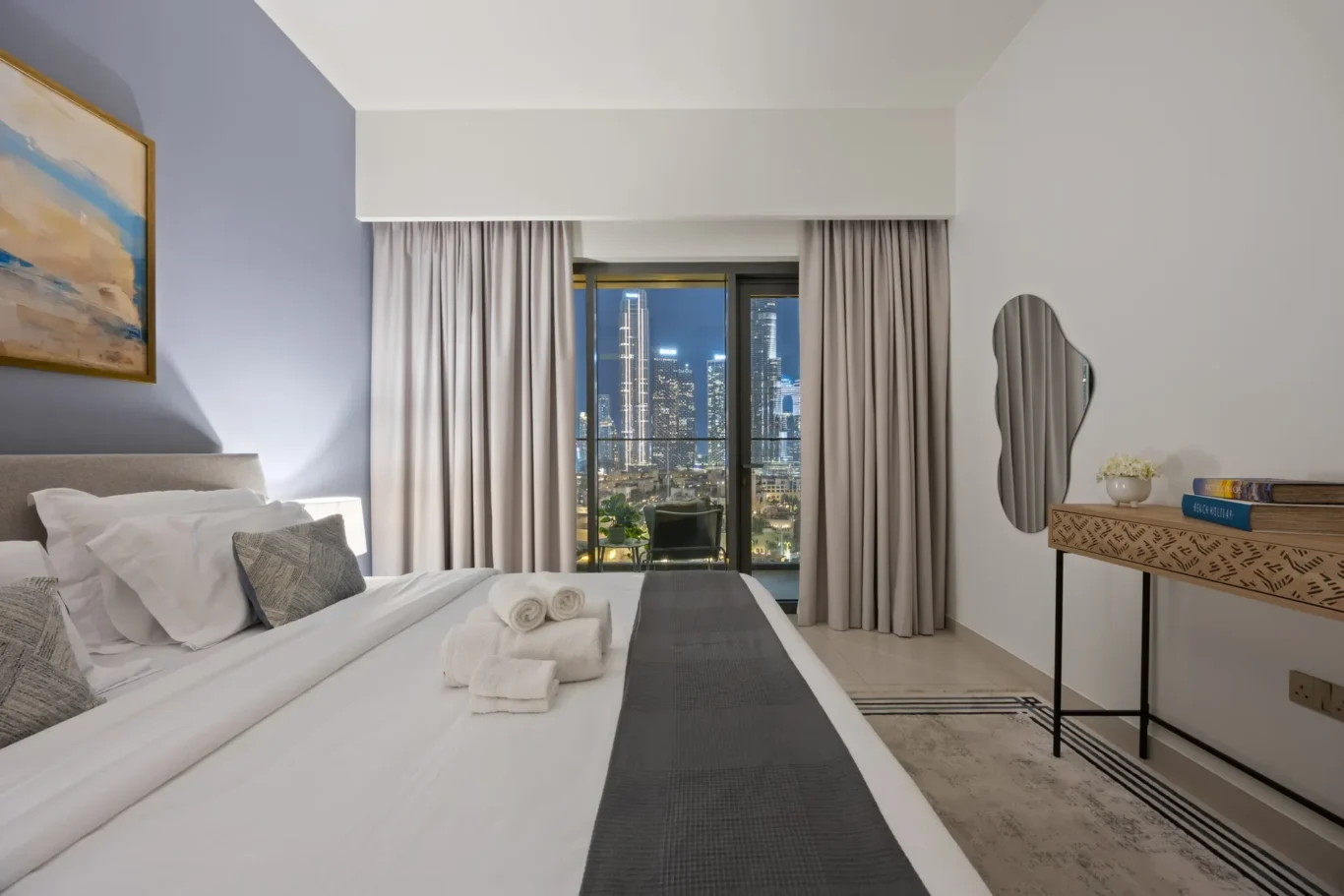 Hotel bedroom with a white bed, gray headboard, and folded towels on the foot; city lights visible through a large window with beige curtains.
