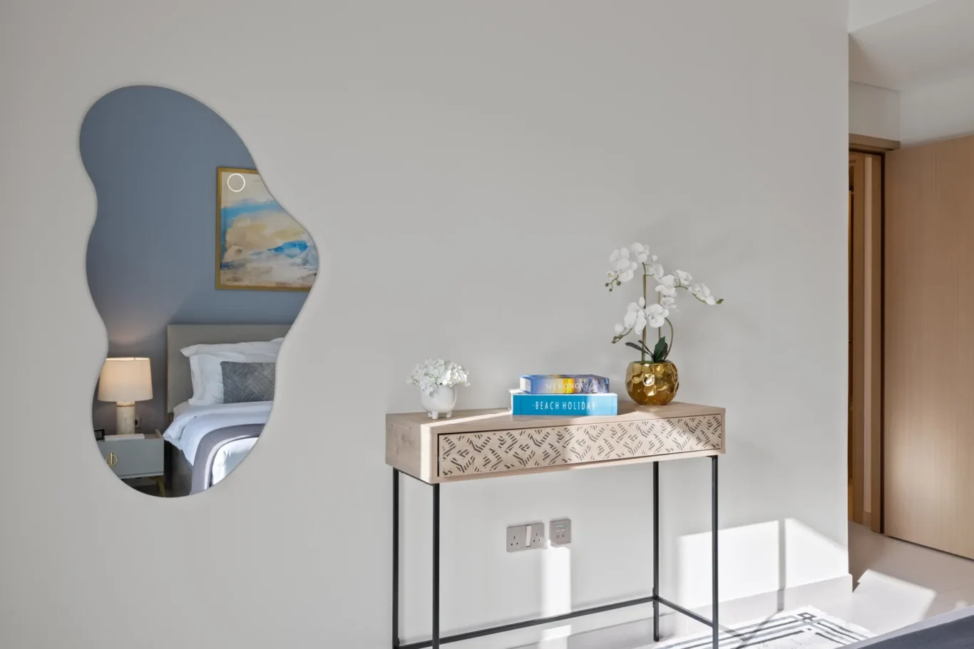 Modern bedroom scene with a kidney-shaped wall mirror reflecting the bed, bedside lamp, and a wooden console with books and white orchids on a pale wall.