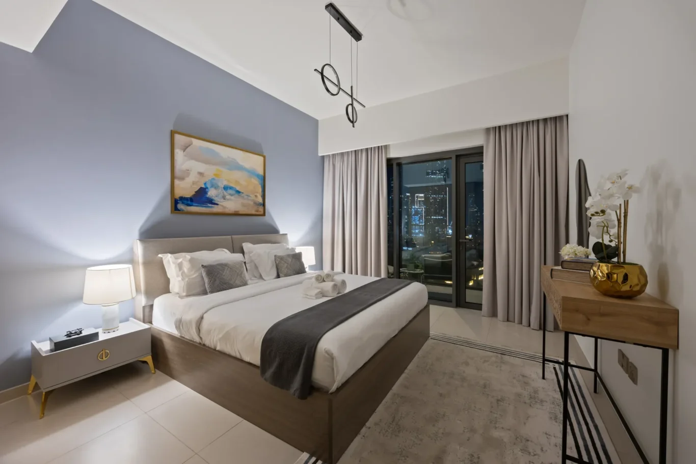 King bed in a modern hotel room with a blue accent wall, abstract painting, and two white nightstands with lamps; city lights visible through glass balcony doors.",