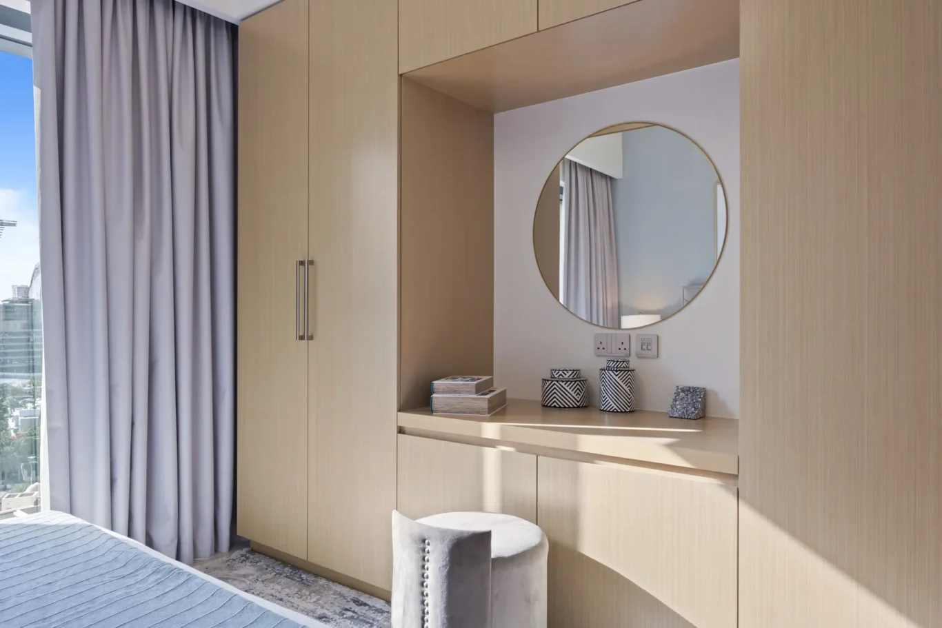 Built-in light wood wardrobe with a round wall mirror, small decorative jars, and a stack of books on a shelf.