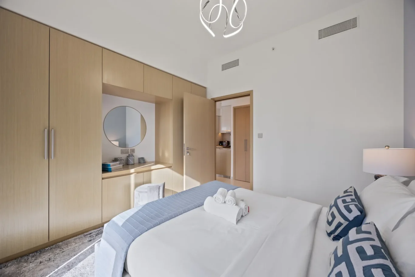 Hotel bedroom with a wall of light wood wardrobes, a desk nook with a round mirror, and a bed with rolled towels on top.