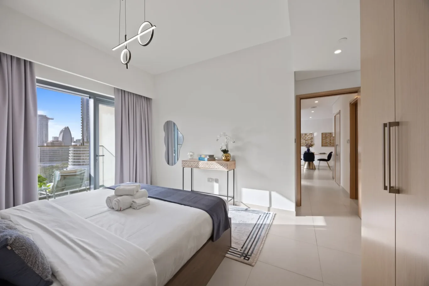 Modern hotel bedroom with two neatly made beds, folded towels on one, and a balcony view of the city skyline.