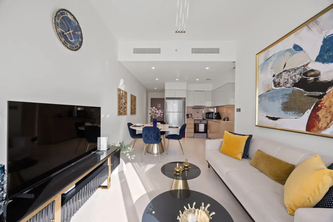 Bright open-plan living area with cream sofa and yellow cushions, round glass coffee tables, and a large abstract painting on the wall to the right.