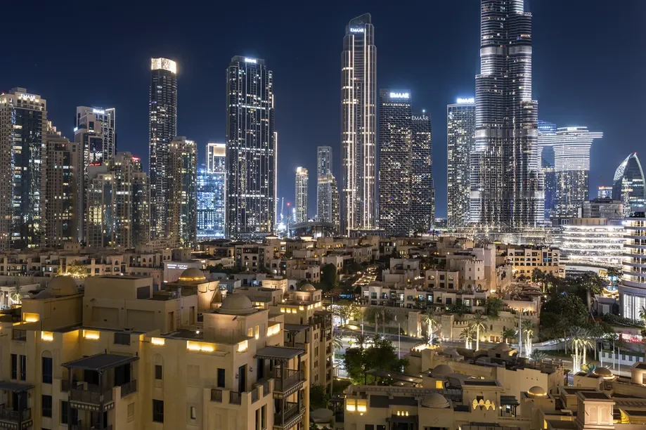 Nighttime cityscape with tall, illuminated skyscrapers rising behind a dense cluster of beige residential buildings in the foreground.