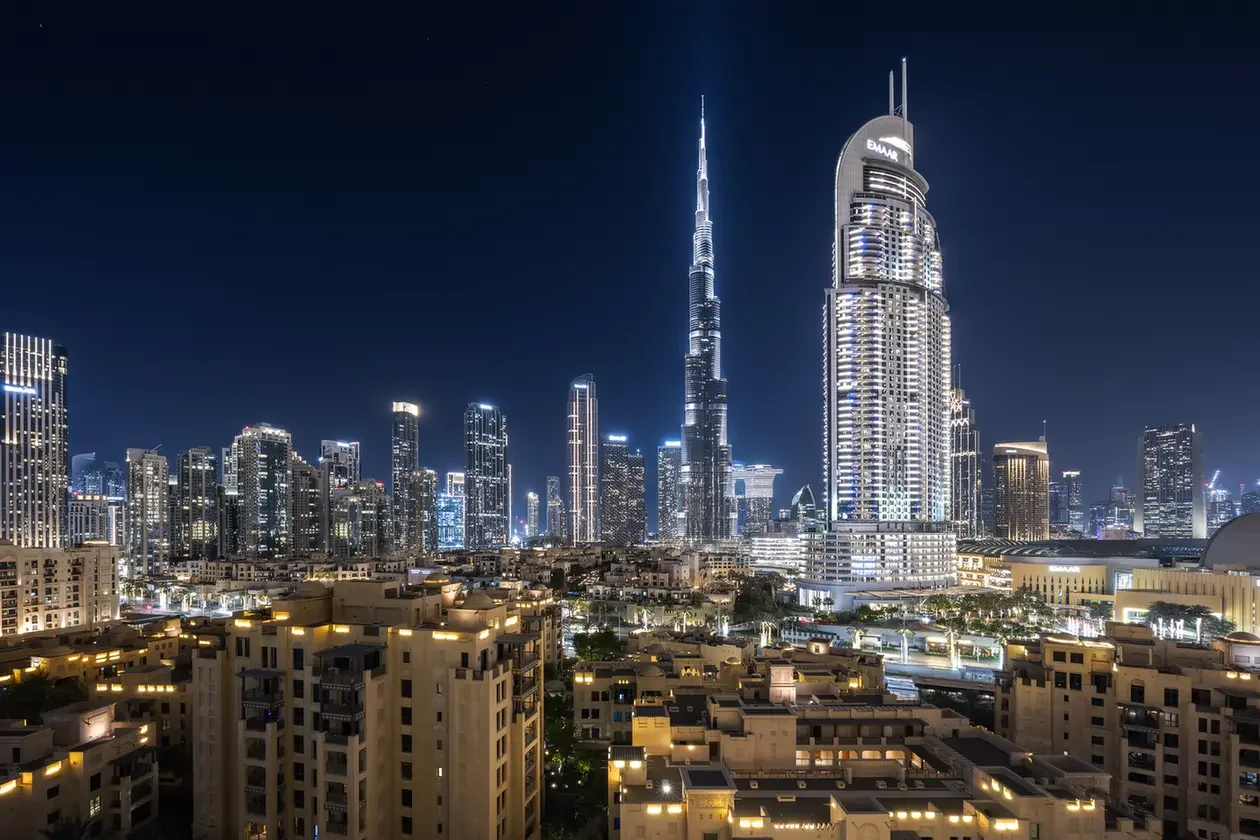 Nighttime city skyline with Burj Khalifa and illuminated skyscrapers over a densely built residential area in the foreground.