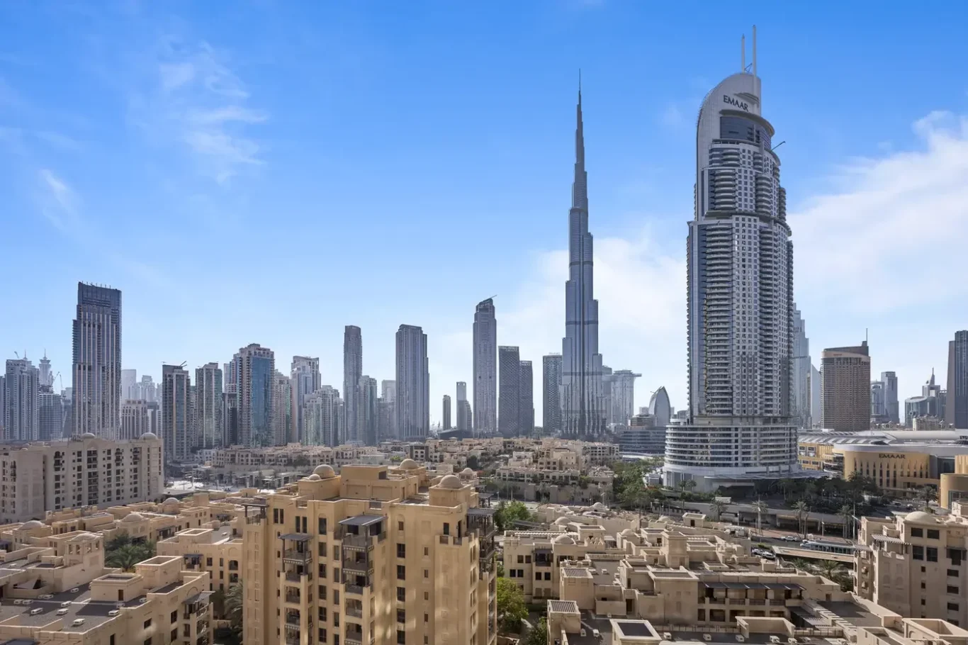 Dubai skyline with Burj Khalifa and curved Emaar tower, beige low-rise foreground under blue sky.