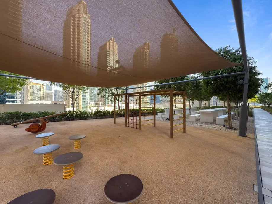 Rooftop playground with a large fabric shade, climbing frame, and round spring seats over a sandy surface with city buildings in the background.