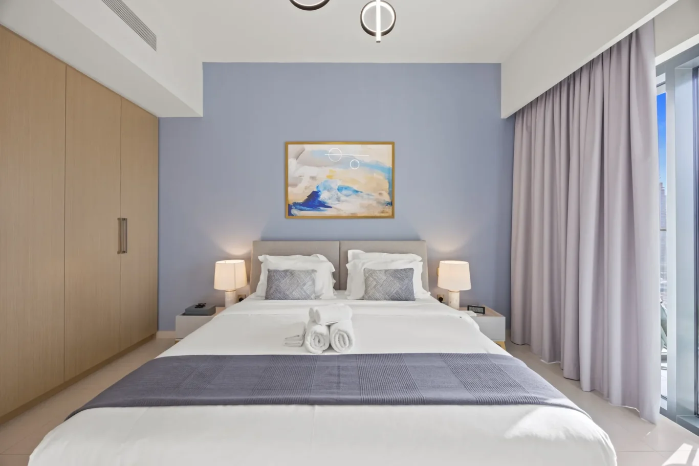 Hotel bedroom with blue accent wall, framed abstract painting above the bed, and two nightstands with lamps