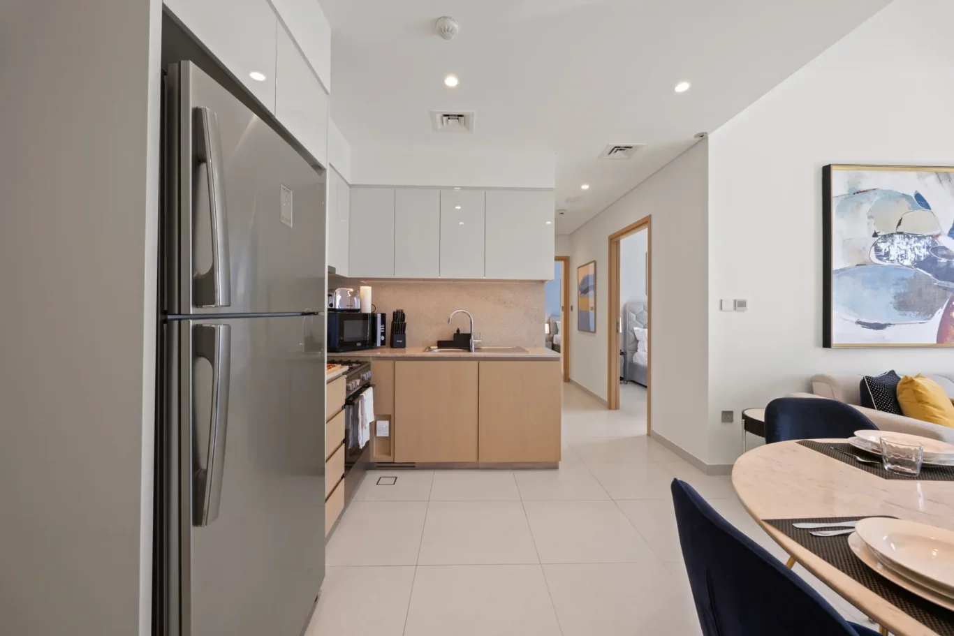 Kitchen with stainless steel double-door refrigerator, white glossy upper cabinets, beige lower cabinets, and a dining table set nearby in a bright open-plan space