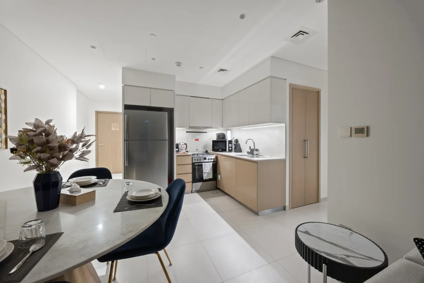 Open-plan kitchen and dining area with a round marble table set for four, blue chairs, and a stainless steel fridge.