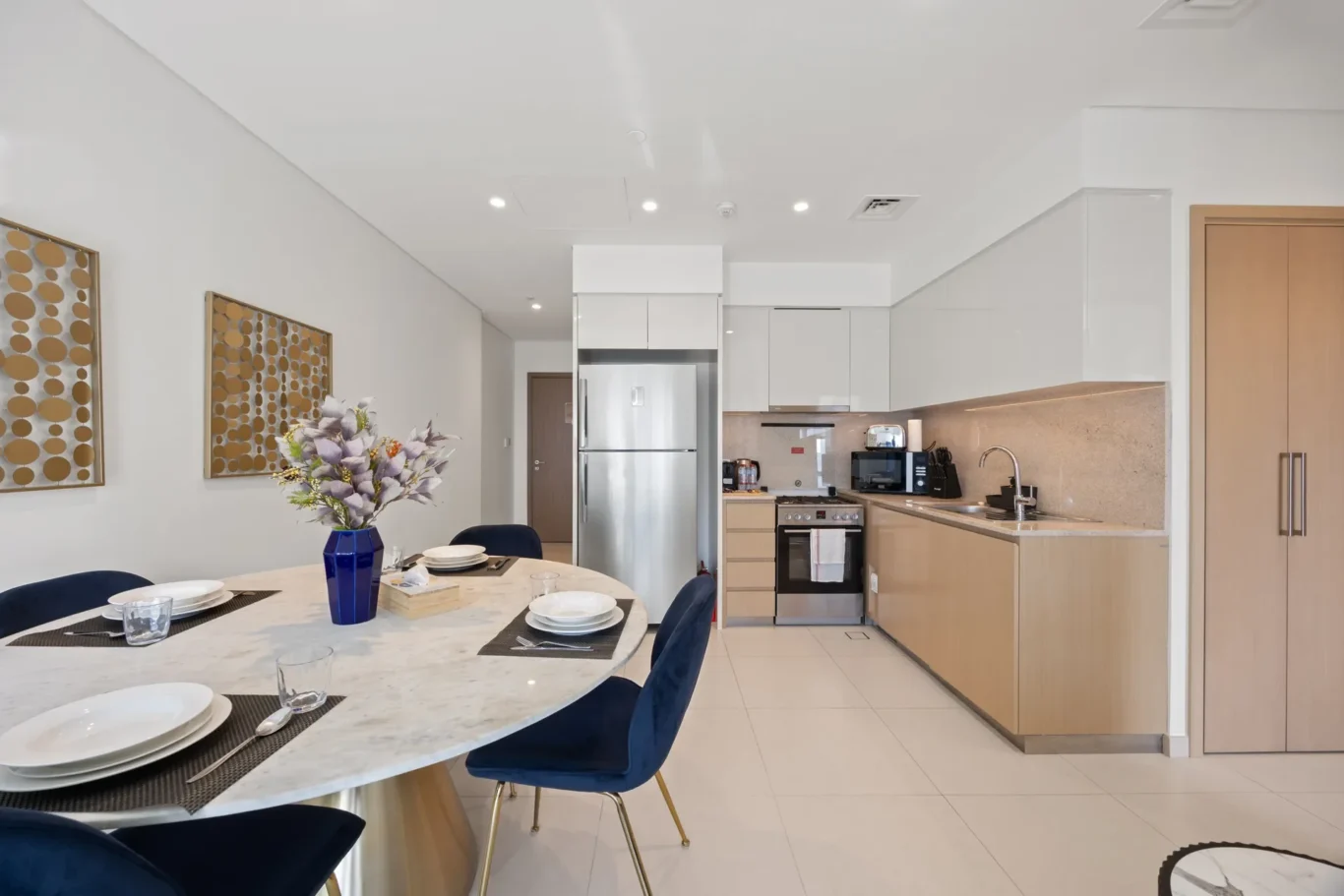 Modern open-plan kitchen with a marble dining island and blue upholstered chairs set for a meal, floral centerpiece visible.