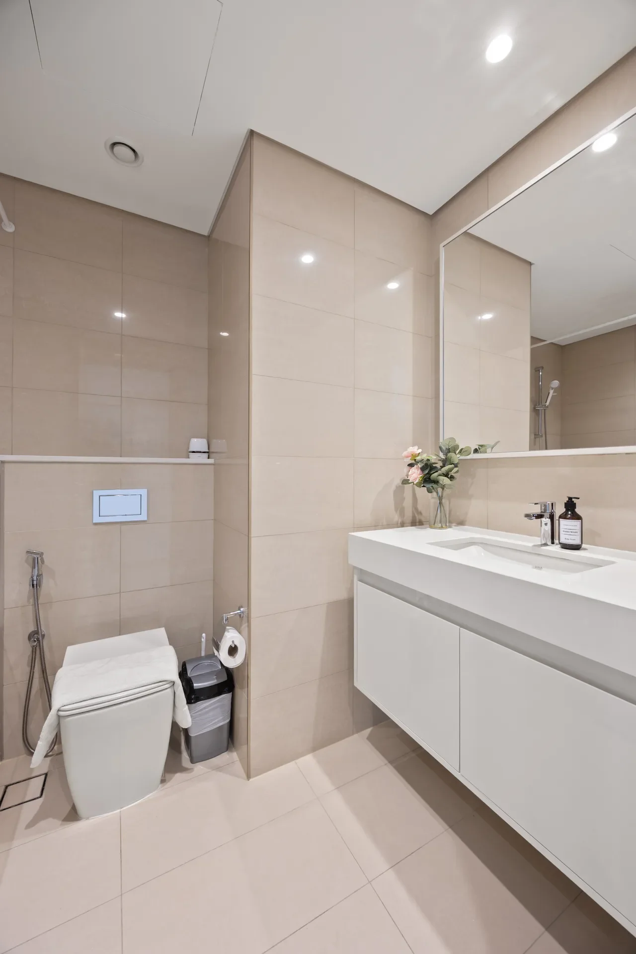 Modern beige-tiled bathroom with a white floating vanity, large mirror, sink, soap bottle, and a small vase of flowers; toilet with bidet spray in the corner. Also shows a shower area in the background.