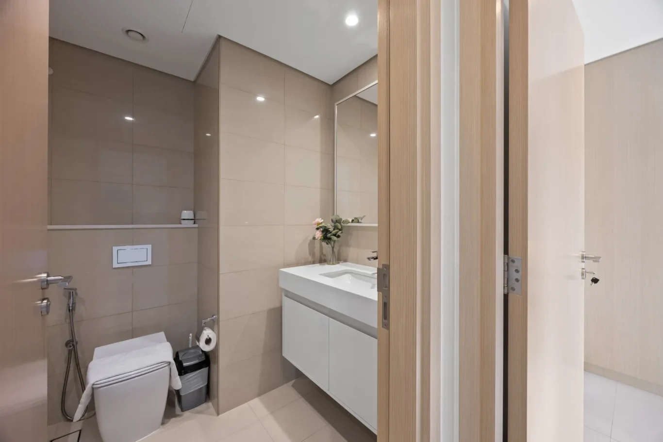 Modern bathroom with beige tile walls, a white vanity with integrated sink, large mirror, and wooden doors nearby.