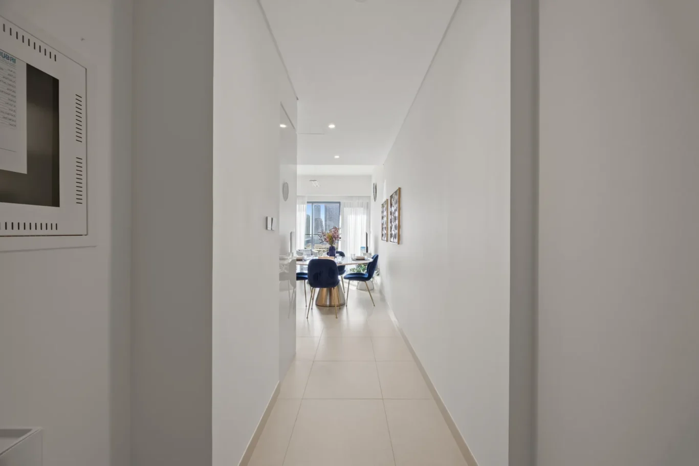 Bright hallway leading to a dining area with a round glass table and blue chairs, city skyline visible through the window on the far end.
