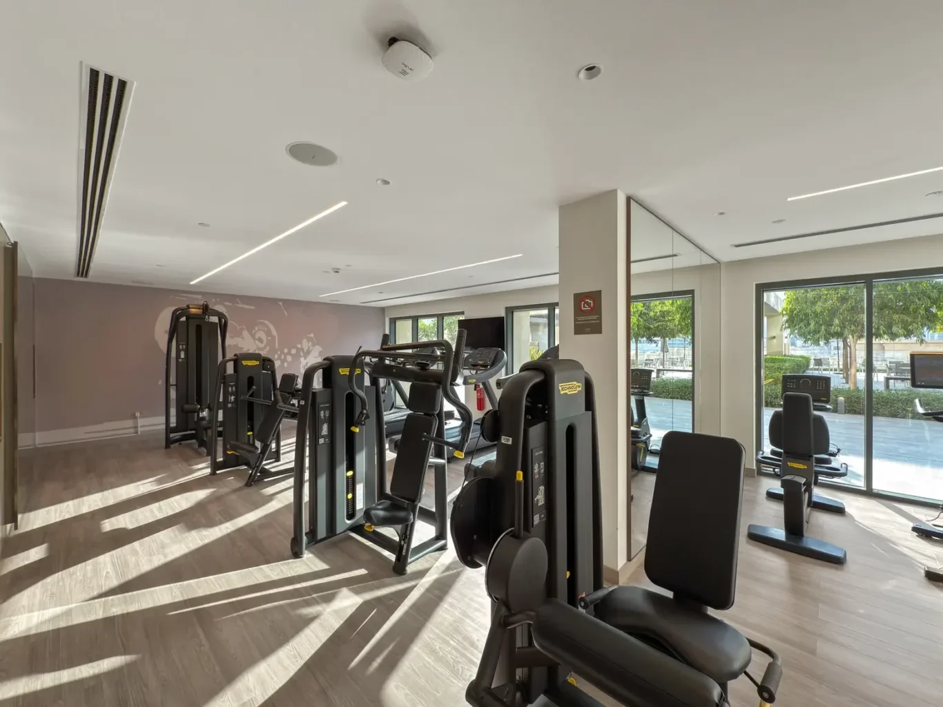 Modern gym with multiple black weight machines and treadmills along a large window wall; wood-like flooring and natural light.
