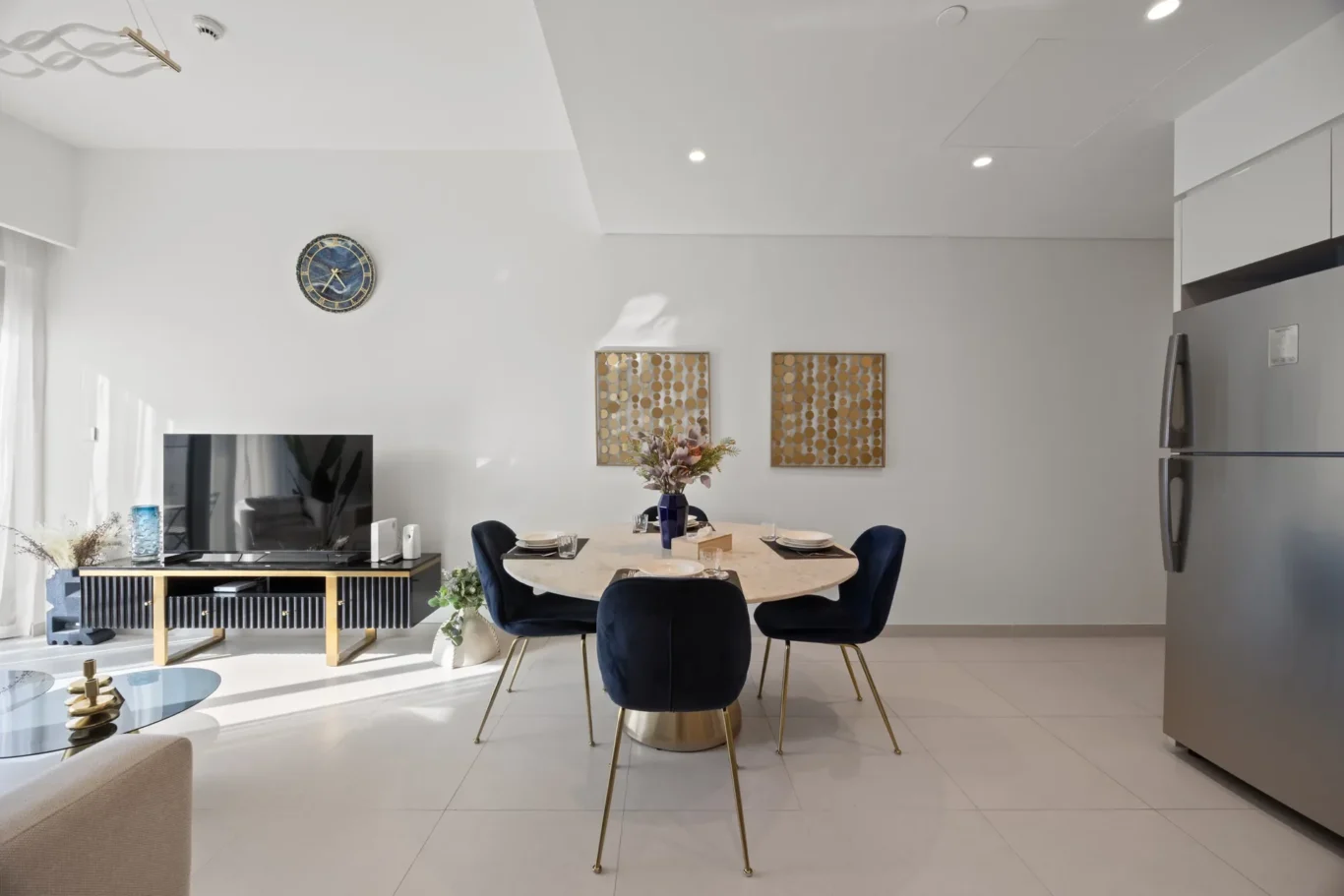 Open-plan dining area with a round marble table set for six, navy chairs, and a bright kitchen with a stainless fridge on the right.