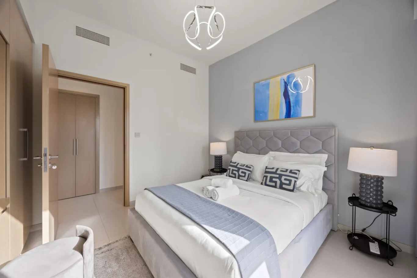 Hotel bedroom with two neatly made beds, gray headboard, and matching throw runners; abstract wall art above the bed and two bedside lamps on round black tables.