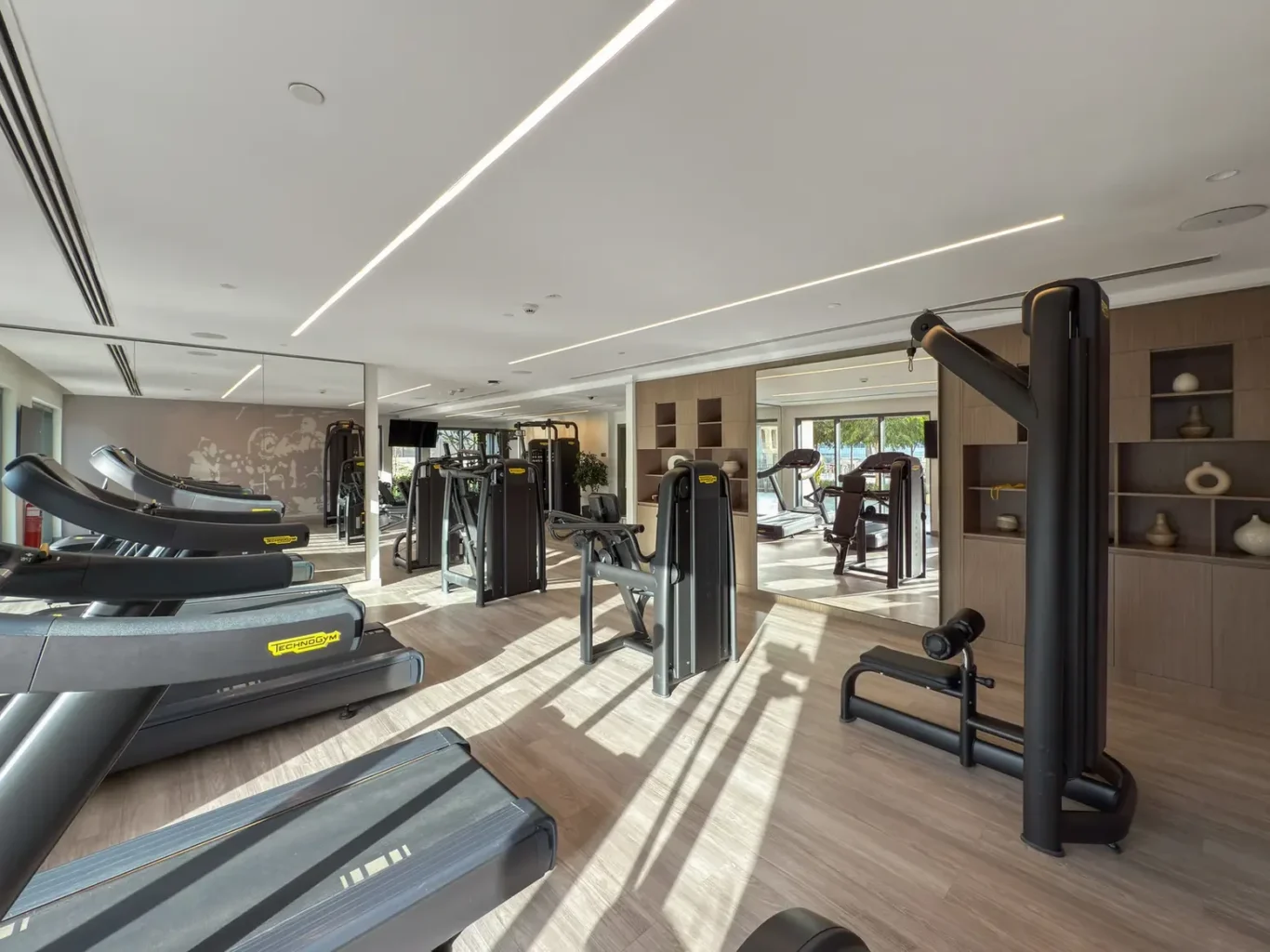 Bright, modern gym with a row of treadmills in the foreground, weight machines in the middle, and large windows letting in natural light.