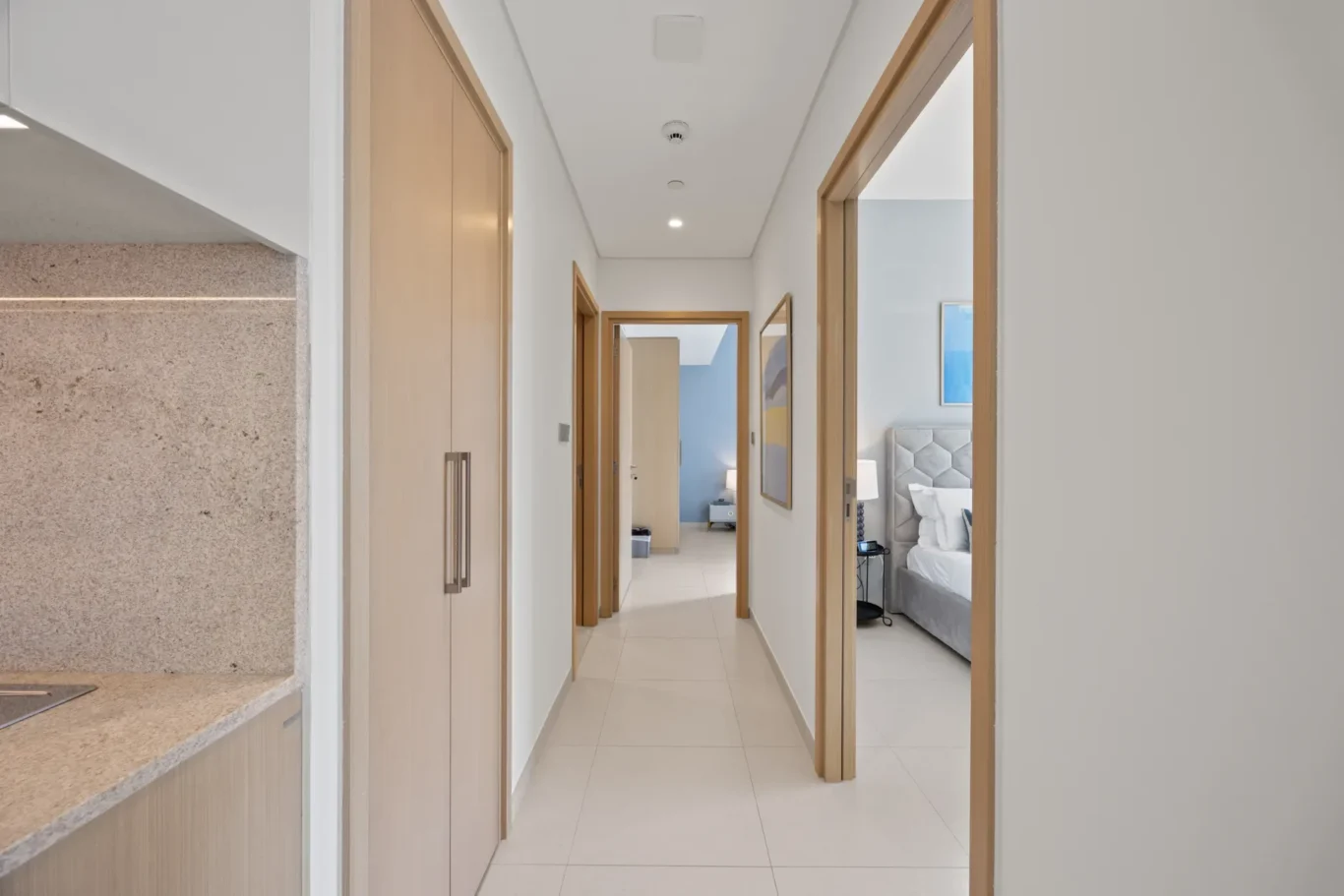 Bright hallway in a modern home with wooden doors on both sides, leading to a bedroom visible at the end with a tufted headboard and blue wall.