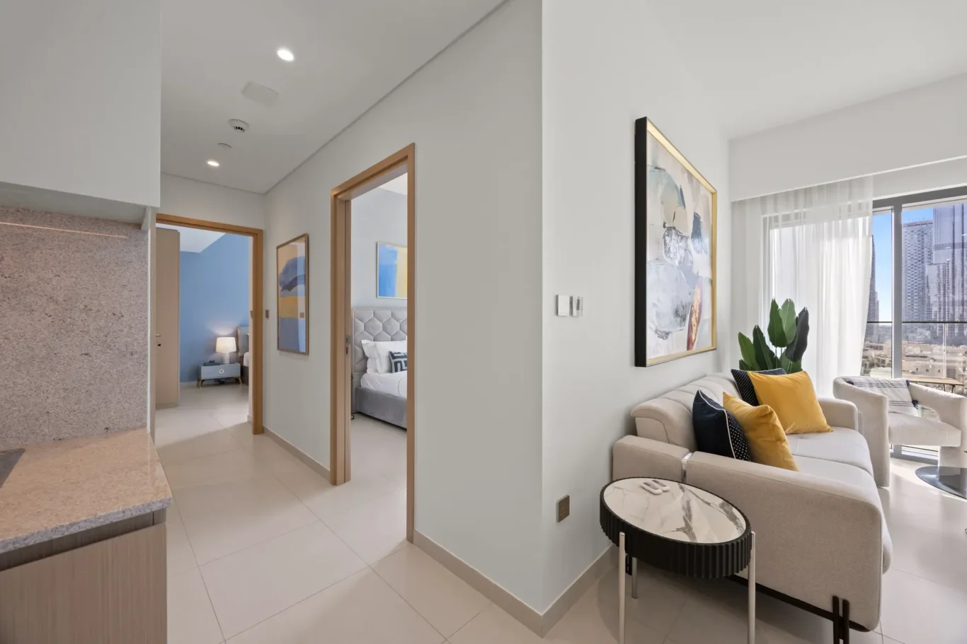 Open-concept living area with beige sofa, yellow cushions, marble-top coffee table, and large window overlooking cityscape.