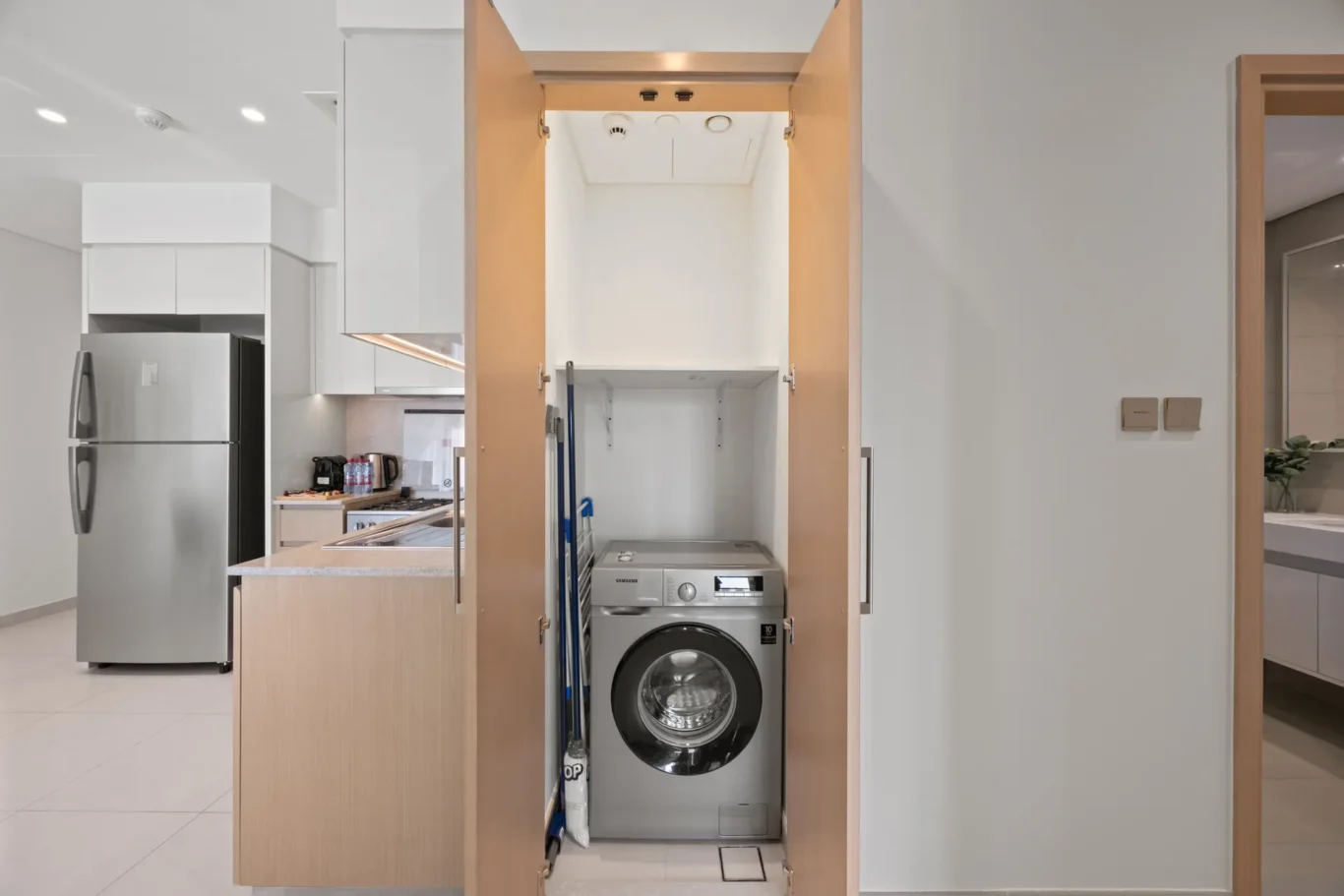 Open cabinet reveals a stacked front-loading washer and dryer in a modern kitchen, with cleaning supplies and a mop nearby.