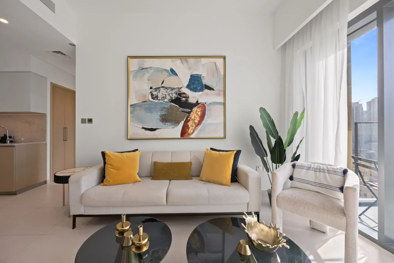 Modern living room with a beige sofa, yellow cushions, and abstract framed artwork above; tall green plant near large windows with sheer white curtains.