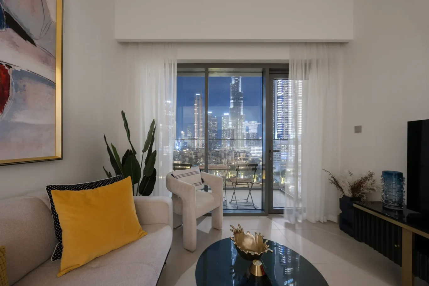 Living room with a beige sofa and yellow cushions, sheer white curtains, and a glass round coffee table; city lights visible through glass balcony doors.