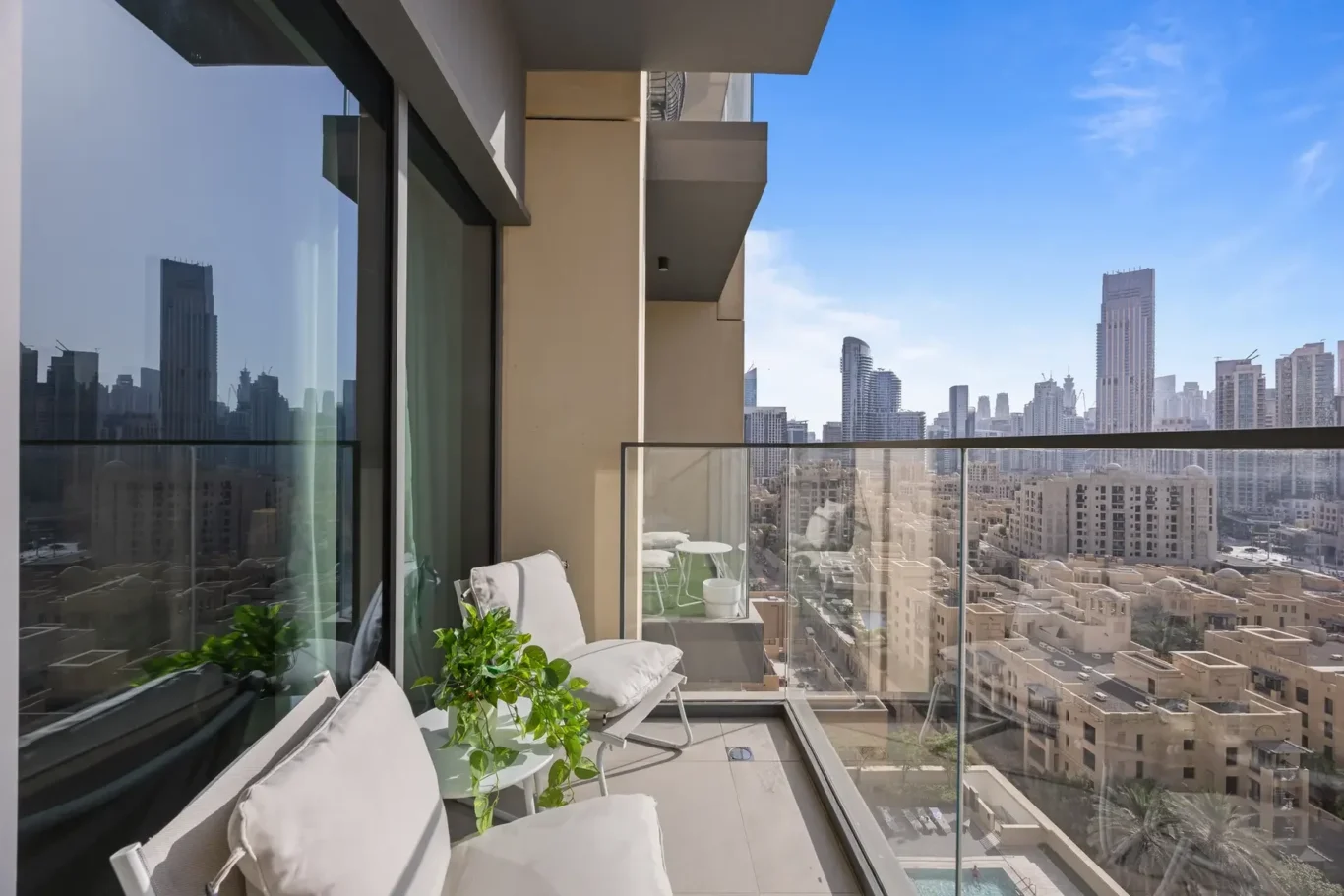 Balcony seating area with white cushioned chairs and a potted plant, glass railing, overlooking a dense city skyline and blue sky.