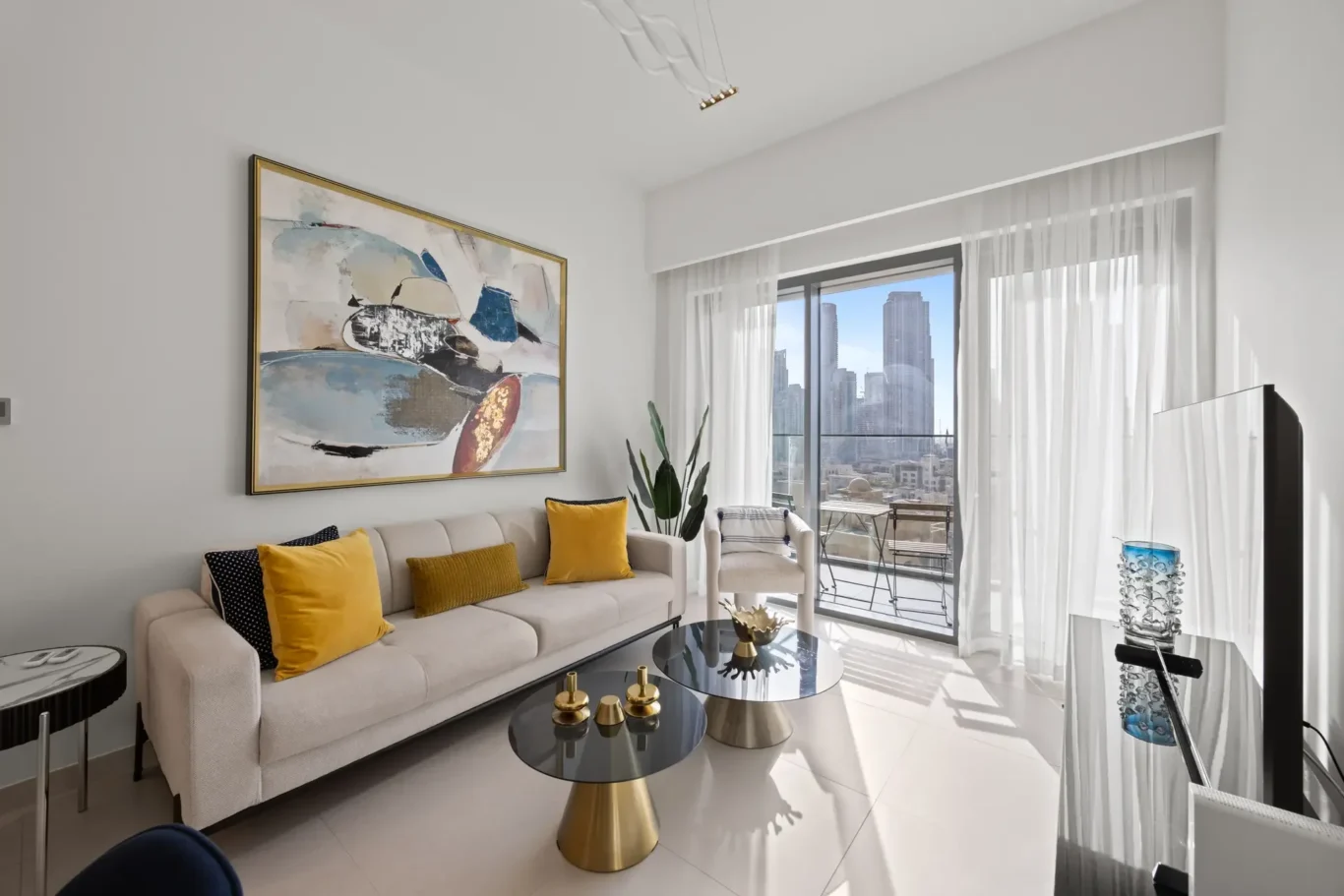 Modern living room with beige sofa and mustard-yellow cushions, abstract wall art, glass coffee tables with gold bases, and a balcony overlooking a city skyline.
