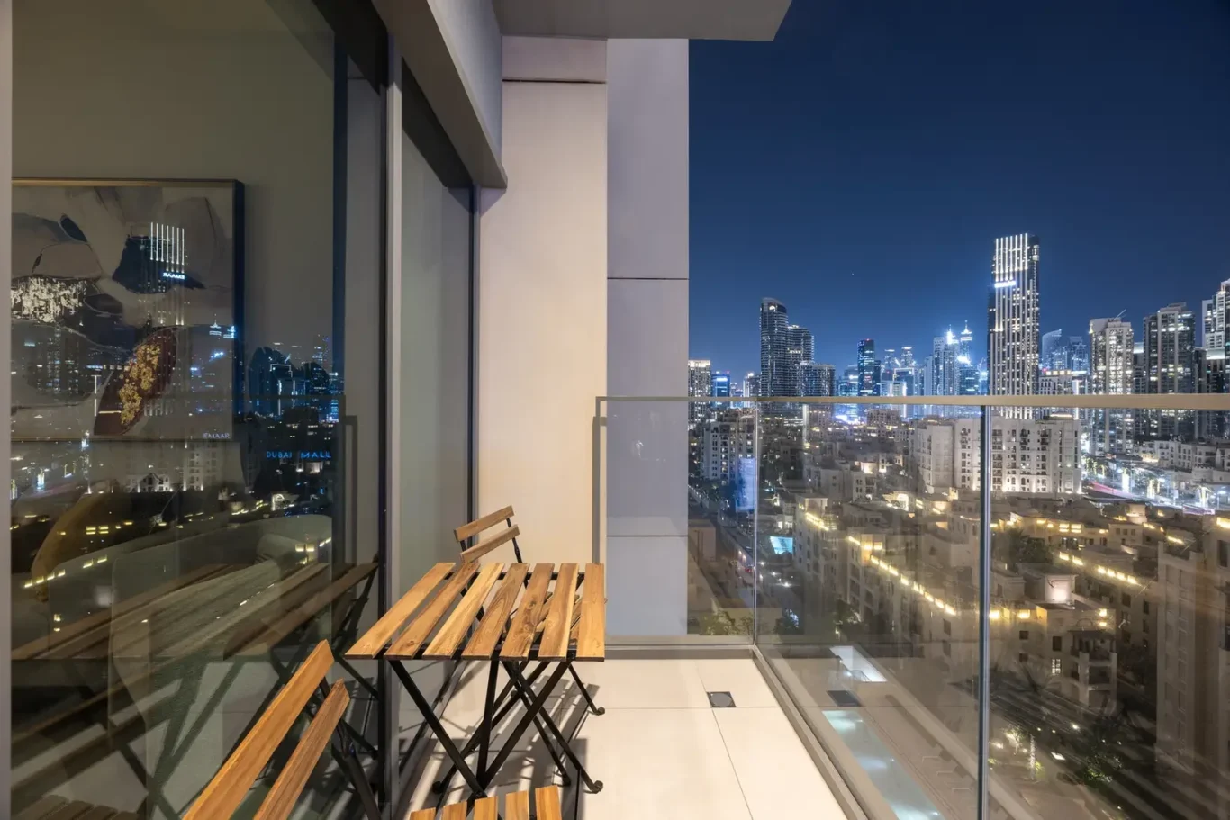 Nighttime city skyline viewed from a balcony, with a small wooden bistro table and chairs in the foreground and a glass railing across the scene.