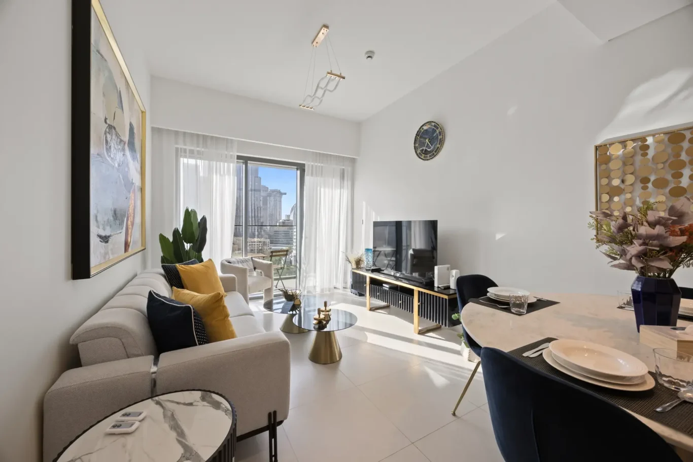 Bright open-plan living room with a light sofa, yellow cushions, marble-top coffee tables, and large windows showing a cityscape outside.