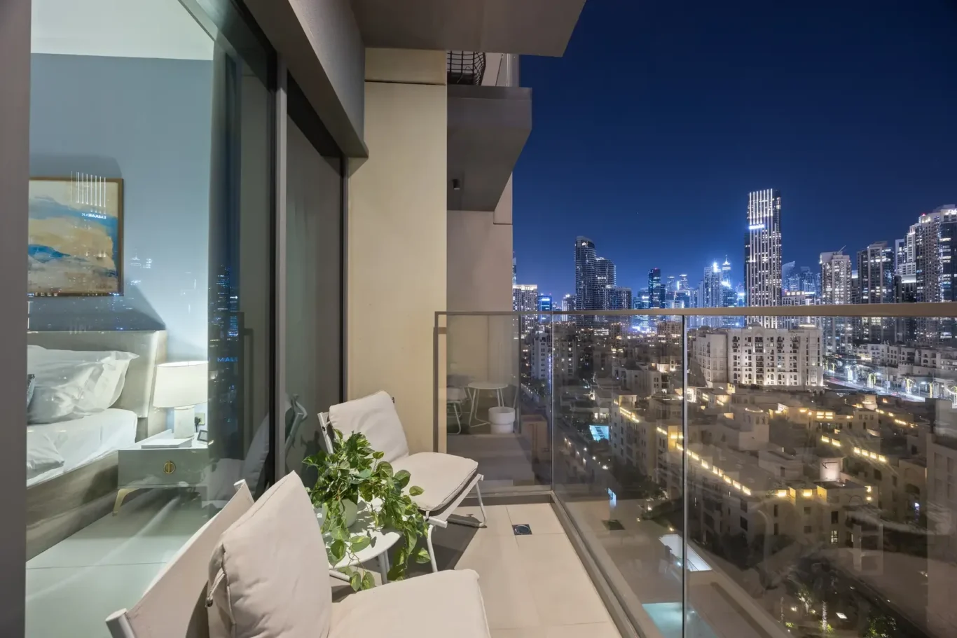 Nighttime city skyline viewed from a glass-walled balcony with lounge chairs and a green plant in the foreground