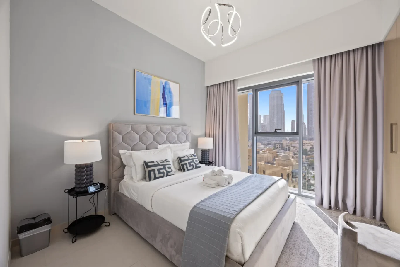Hotel bedroom with a tufted gray headboard, white bedding, two nightstands, and a large window showing a city skyline.