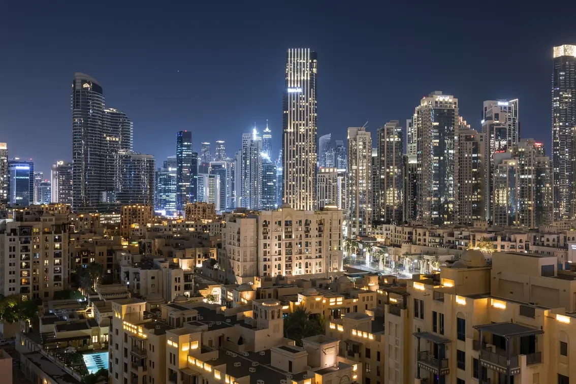 Nighttime city skyline with tall glass skyscrapers and warm-lit residential buildings in the foreground.