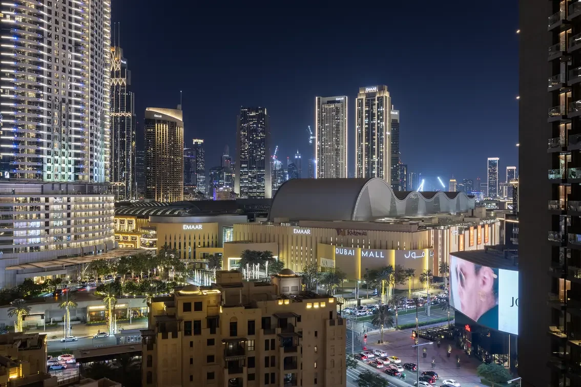Nighttime cityscape of Downtown Dubai with the Dubai Mall complex, Emaar signage, and tall illuminated skyscrapers in the background.