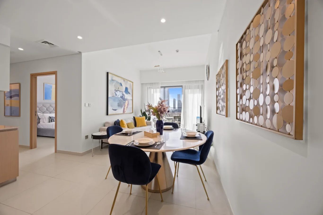 Open dining area with a round glass table, blue chairs, and a city skyline view through large windows/doors on the right side of the room.