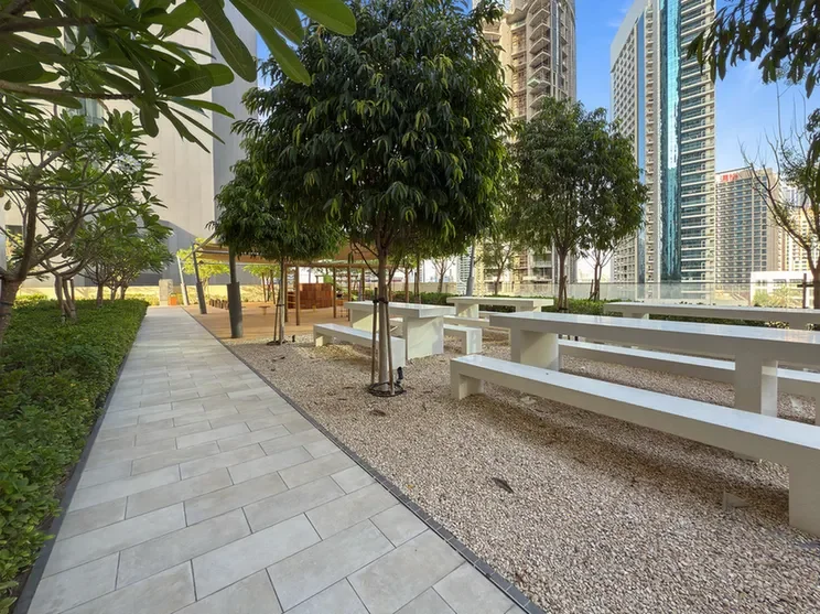 Urban park path with trees, picnic tables, and long white benches; high-rise buildings in the background.