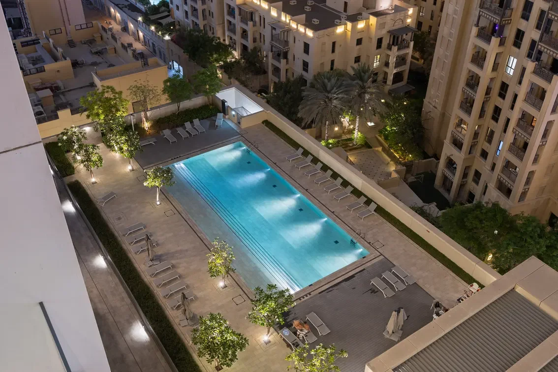 Night aerial view of a rectangular pool in a residential complex, with lounge chairs, trees, and surrounding apartment buildings at dusk.