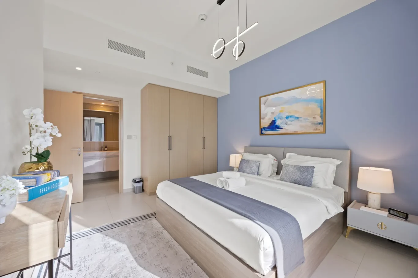 Hotel bedroom with two twin beds side by side, blue accent wall, abstract painting, and a wooden wardrobe; door to a bright bathroom visible in the background.