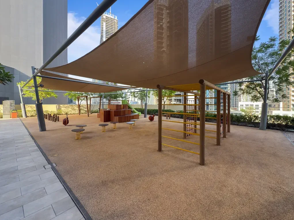 Outdoor playground under large shade sails in a city courtyard, with climbing bars, stepping stools and wooden seating nearby.