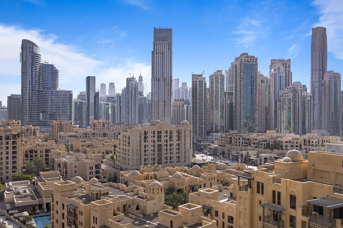 Skyline of tall glass skyscrapers rising behind a foreground of beige desert-style apartment buildings under a blue sky.