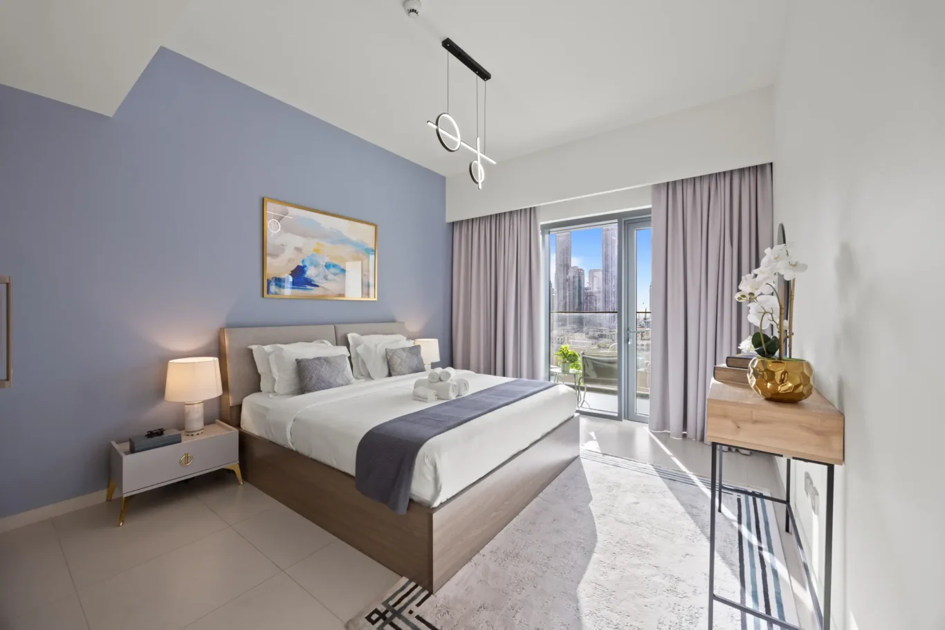 Modern hotel-style bedroom with a blue feature wall, large bed with white linens, gray throw, and two bedside tables. City-view balcony doors are open to skyscrapers outside.