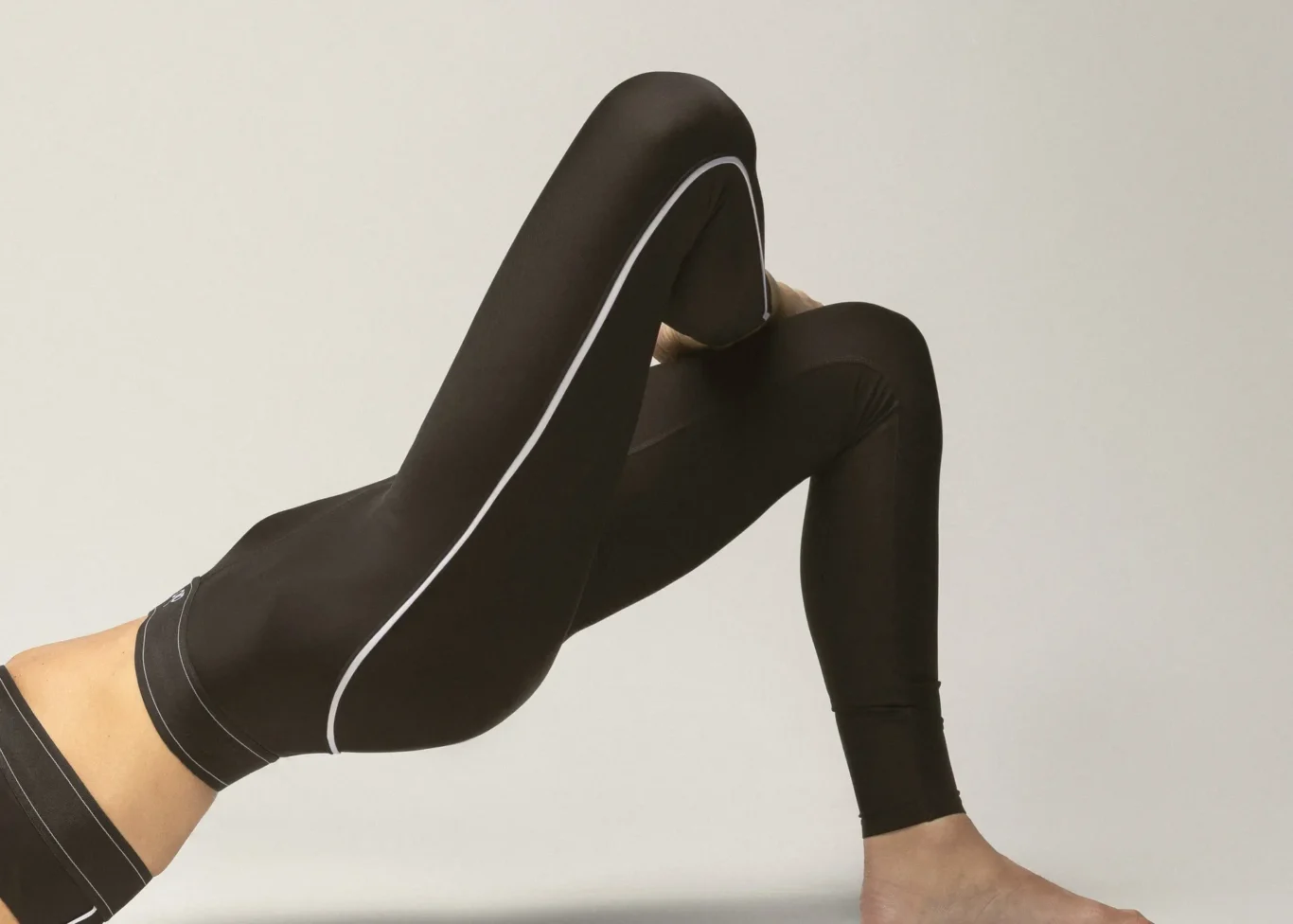 Person wearing black athletic leggings with white piping, bending one leg in a stretch against a neutral background.
