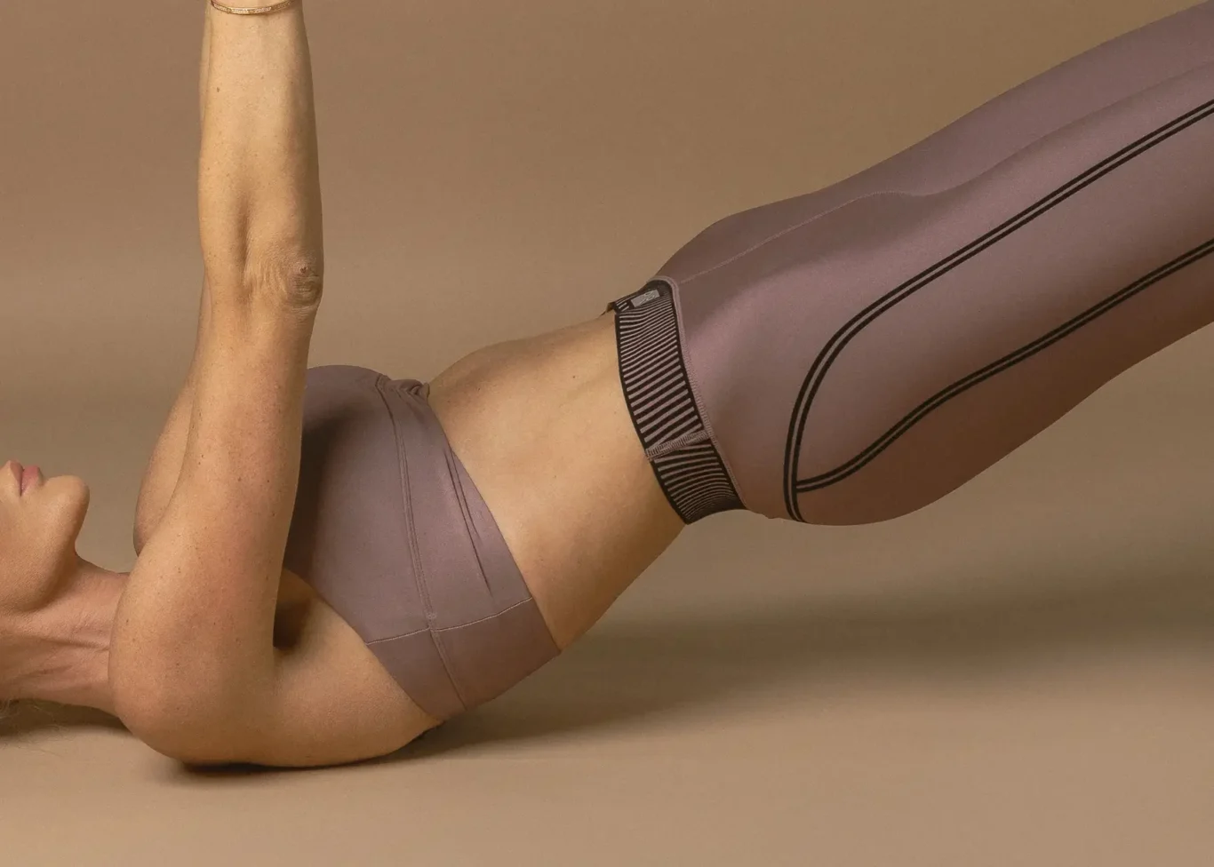Person wearing mauve activewear lying on their back with legs raised and arms extended upward, performing a core exercise