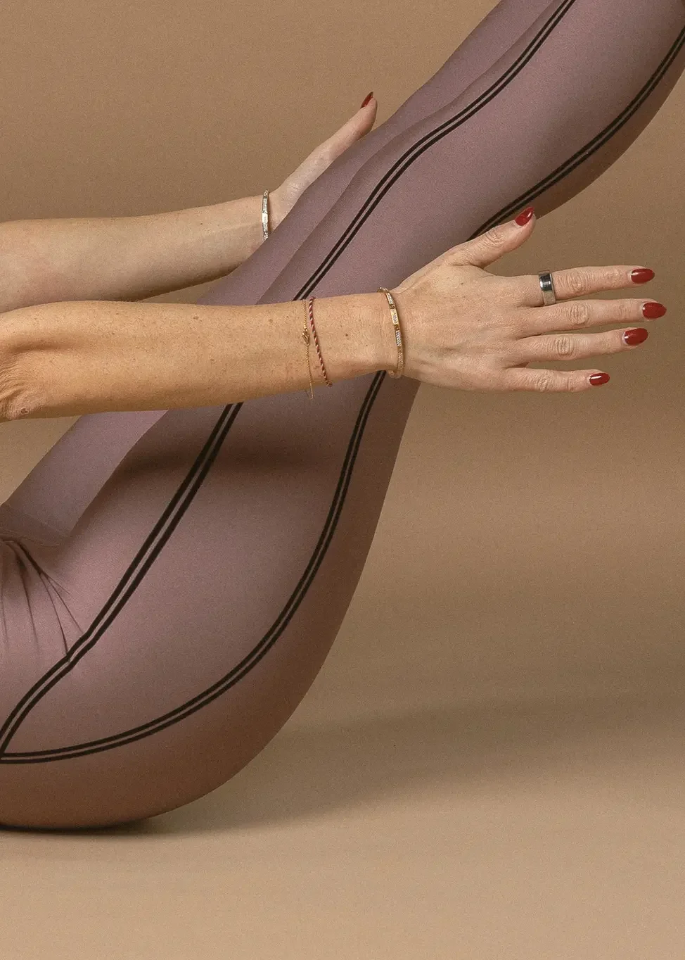 Person in mauve athletic leggings with black stripes stretches their legs, holding ankles with hands showing red nail polish and bracelets.