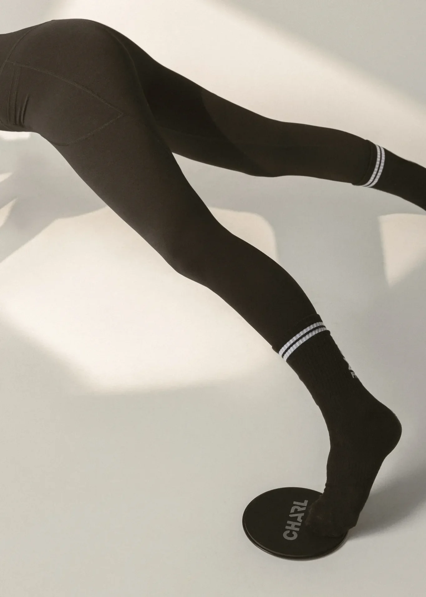 Close-up of a person in black leggings performing a balance exercise on a round black balance disc on a white floor.