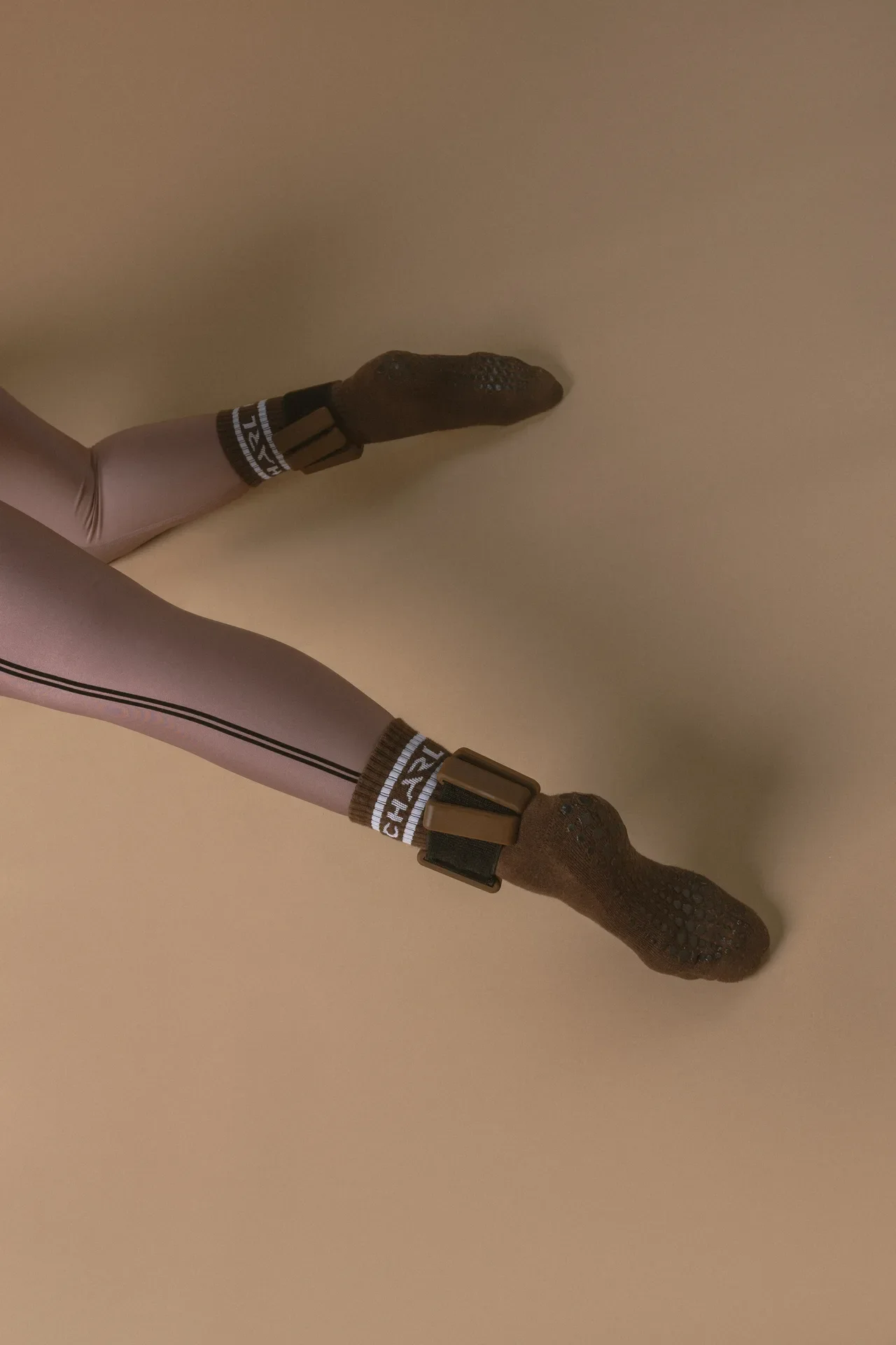 Two legs extended on a beige surface, wearing lavender tights with a dark brown sock-like foot covering and white-trimmed ankle straps.