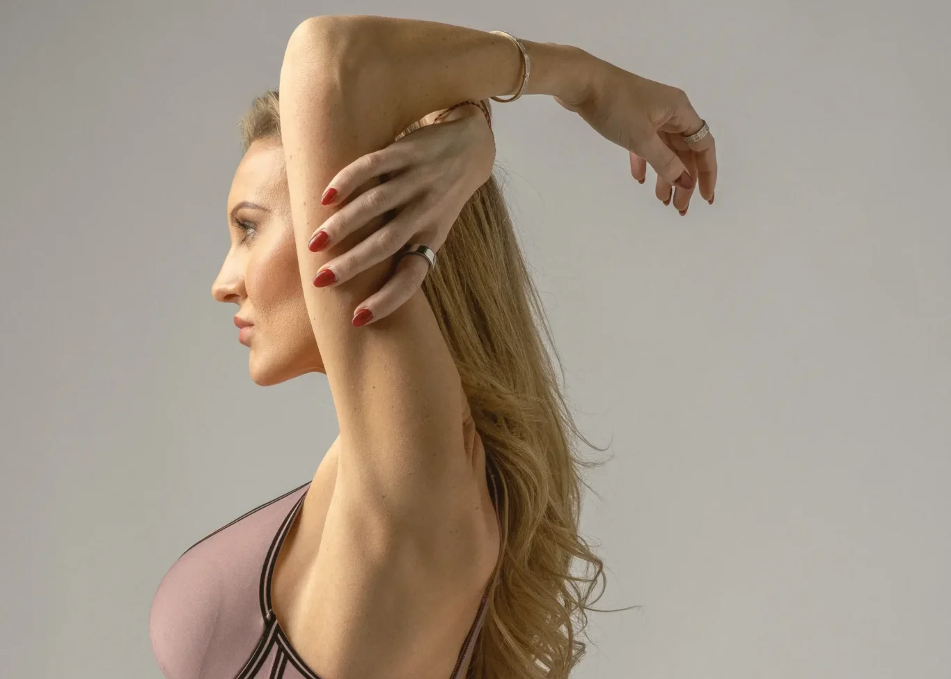 Side profile of a woman with arms raised, wearing a blush-pink athletic top; red nails and rings visible against a gray background.