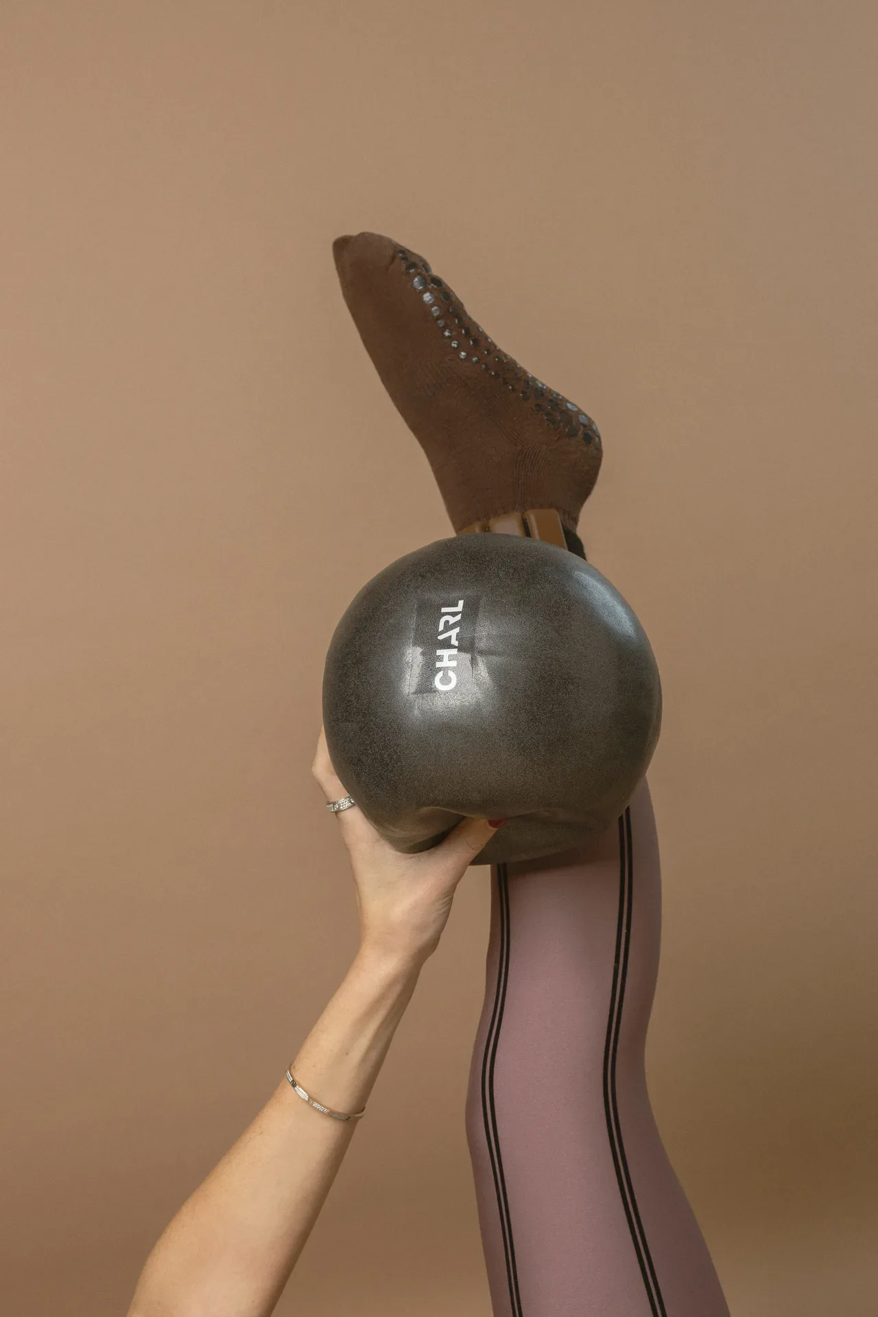 Person holding a black exercise ball against a brown wall with a leg raised in mauve tights above the ball.